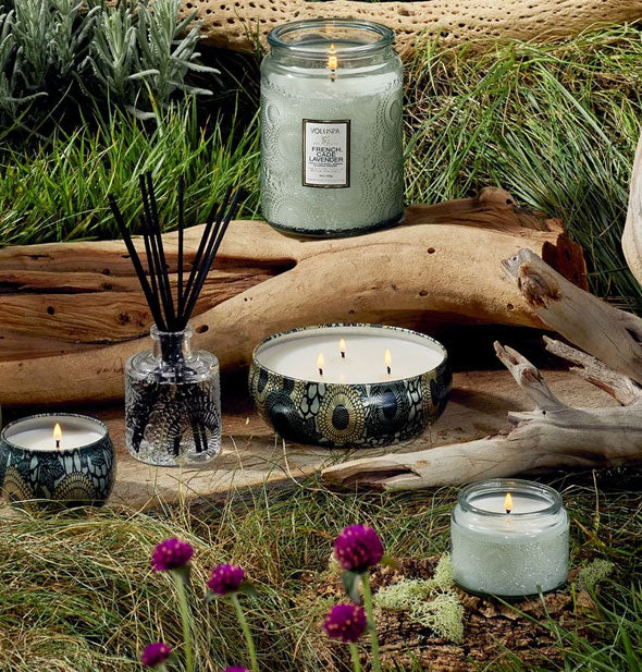 Assortment of Voluspa candles and a reed diffuser sit among driftwood and purple wildflowers on a grassy backdrop