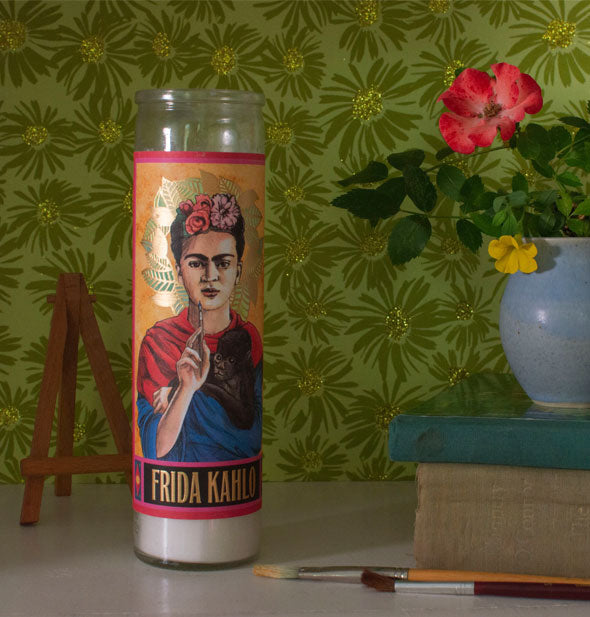 Frida Kahlo prayer candle on a tabletop with flower vase, paint brushes, and tiny easel against sparkly green floral wallpaper