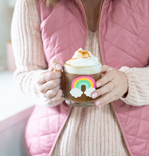 Model poses holding a clear glass Fuck Mornings rainbow mug filled with a frothy beverage inside
