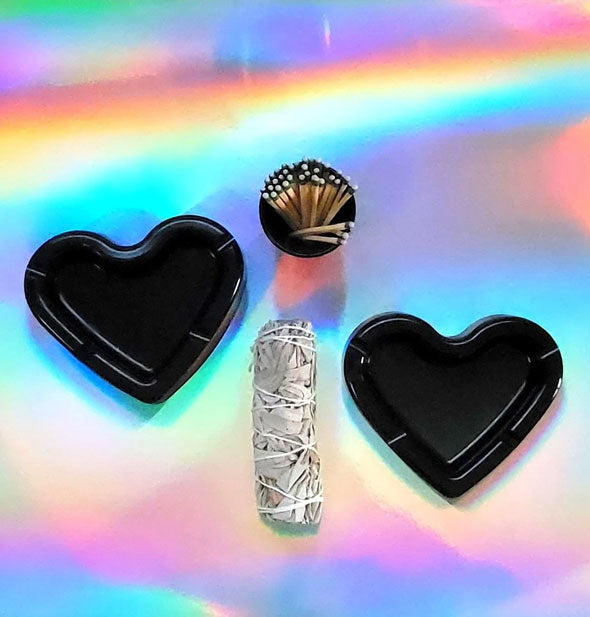 Two black heart-shaped ashtrays staged with matches and a sage bundle on holographic surface