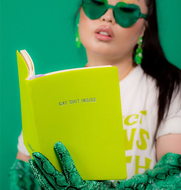 Model wearing green heart-shaped sunglasses, green dangle earrings, and green snake print elbow-length gloves holds a Gay Shit Inside journal open