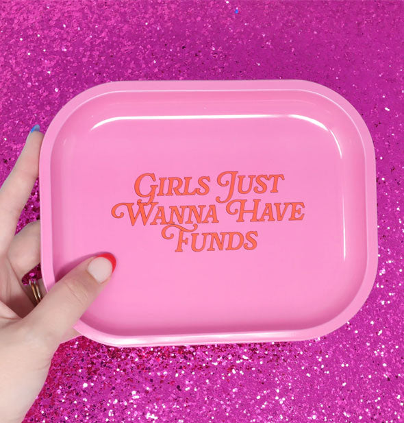 Model's hand holds a pink Girls Just Wanna Have Funds tray against a dark pink glittery backdrop