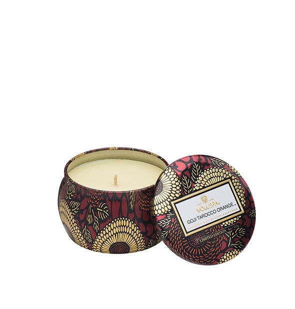 A small unlit candle inside a rounded tin with red and gold metallic floral design and matching lid set to the side.