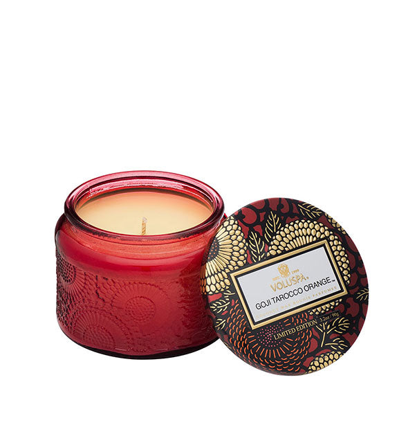 Red embossed glass Goji Tarocco Orange Voluspa candle har with red and gold metallic floral lid set to the side