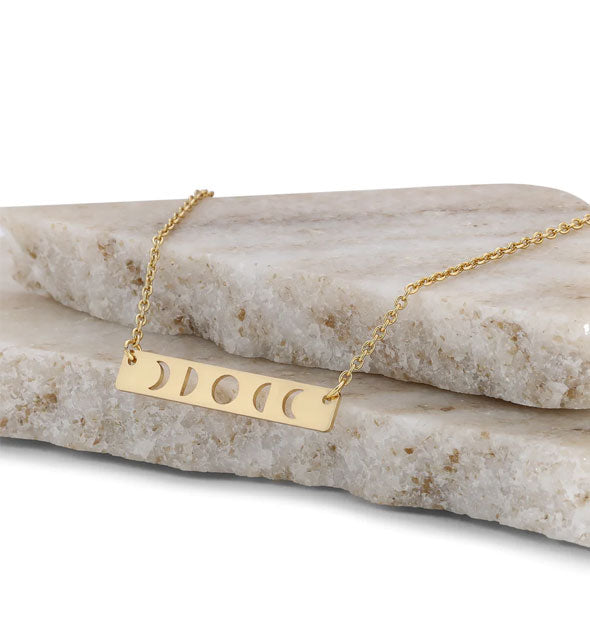 Gold cutout moon phases bar charm necklace rests on two slabs of marble