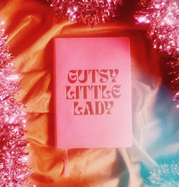 Pink Gutsy Little Lady journal rests on a piece of fabric surrounded by pink tinssel