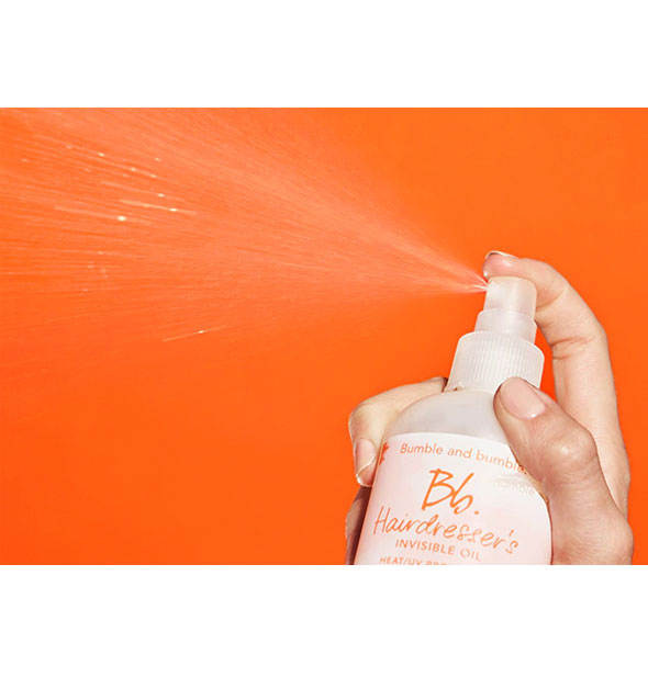 Model's hand dispenses a mist of Bumble and bumble Hairdresser's Invisible Oil Heat/UV Protective Primer against an orange backdrop to show spray pattern