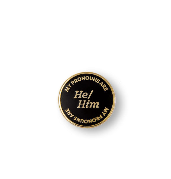 Round black enamel pin with gold border says, "My Pronouns Are" at top and bottom and "He/Him" in the center, all in gold lettering