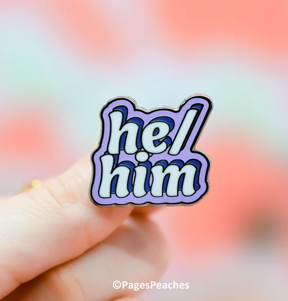A purple enamel He/Him pin is held between a model's fingers with Pages Peaches image copyright at bottom