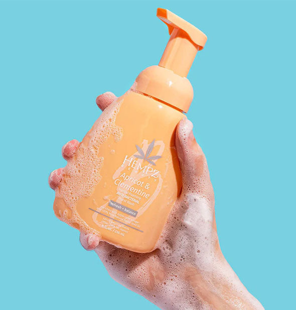 Model's hand covered in soapy suds holds a bottle of Hempz Apricot & Clementine Herbal Foaming Antibacterial Hand Wash up in front of a blue background