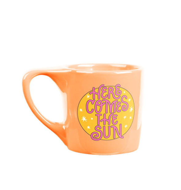 Peach-colored coffee mug with triangular handle says, "Here comes the sun" in pink with a yellow star-patterned circle graphic