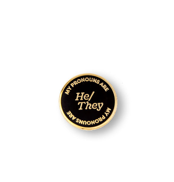Round black enamel pin with gold border says, "My Pronouns Are" at top and bottom and "He/They" in the center, all in gold lettering