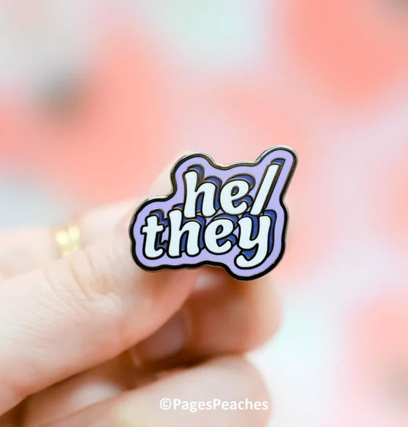A purple enamel He/They pin is held between a model's fingers with Pages Peaches image copyright at bottom