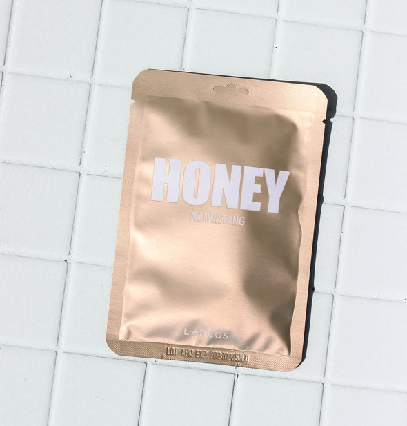 Shiny gold Honey sheet mask packet on a white tiled surface