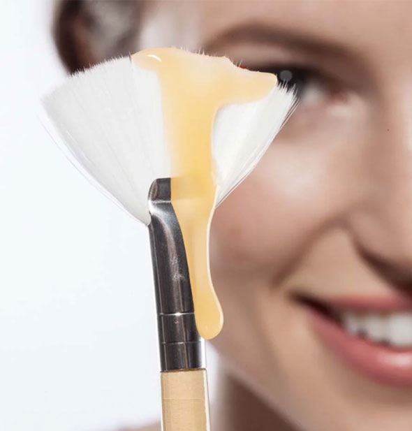 Smiling model holds forward a white fan brush with a dribble of Honey Heel Glaze running from bristles down handle