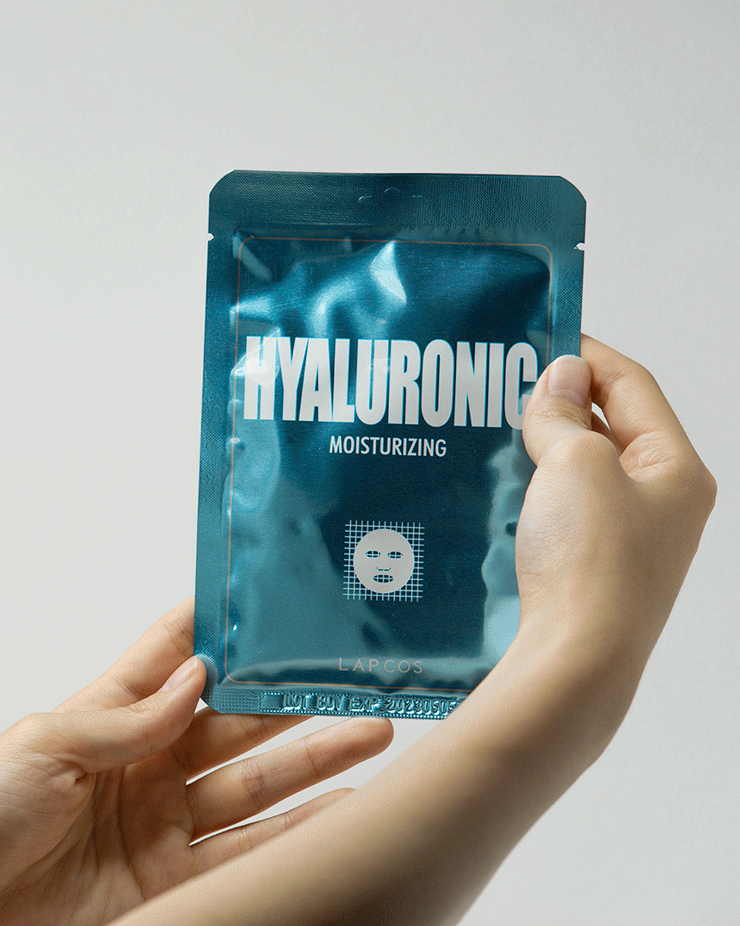 Model's hands open a Hyaluronic Moisturizing sheet mask packet and unfold the mask inside