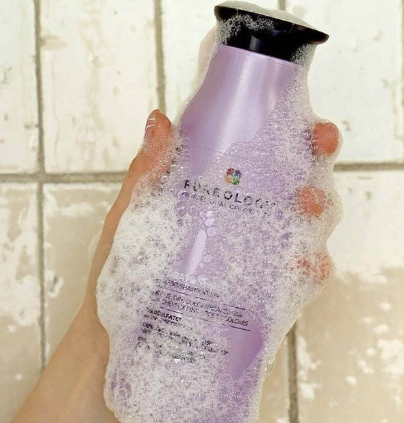 Model's hand holds a bottle of Pureology Hydrate Sheer Shampoo that's covered in lather