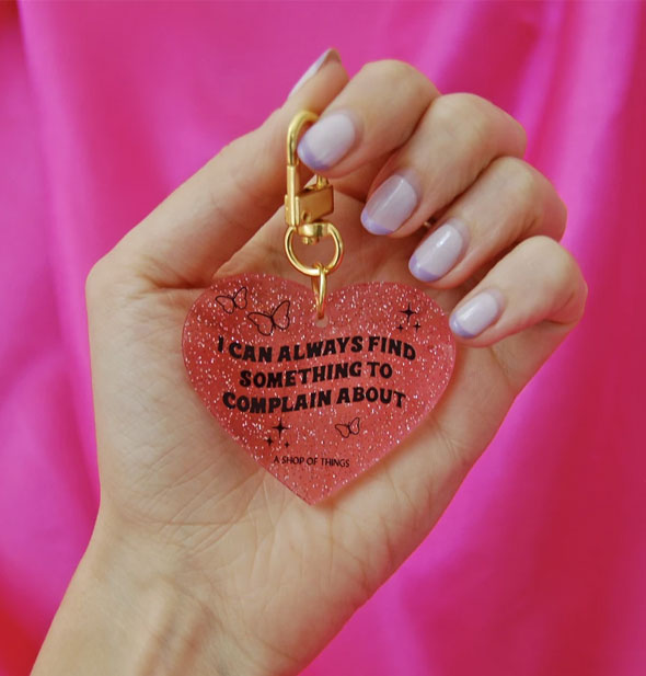 Model's hand holds a pink glitter heart-shaped keychain that says, "I can always find something to complain about" in black lettering accented with butterflies and stars