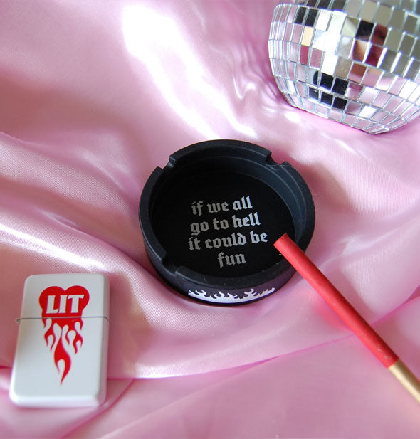 Round black ashtray that says, "If we all go to hell it could be fun" is staged with a cigarette, lighter, and disco ball on pink satin