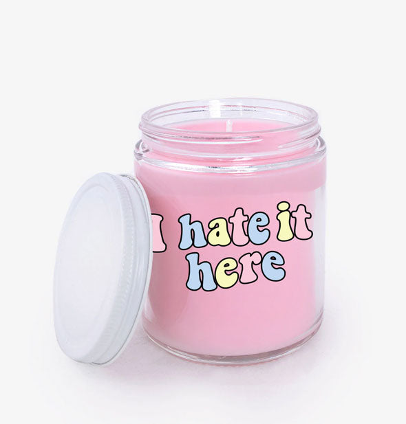 Pink wax jar candle with white lid says, "I hate it here" in a retro-style multicolor font