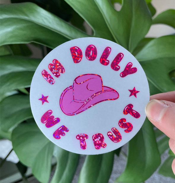 Model's hand holds a round "In Dolly We Trust" sticker with pink glitter bubble lettering and cowgirl hat graphic in the center; green monstera leaves are in the background