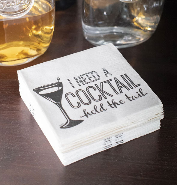 Stack of I Need a Cocktail...Hold the Tail cocktail napkins on a bar with glasses