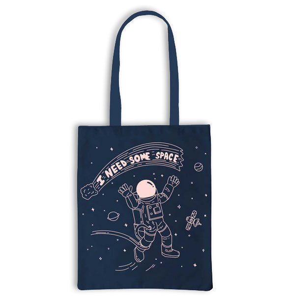 Dark blue tote bag features light pink illustration of an astronaut floating in outer space underneath a comet that says, "I need some space" in its tail
