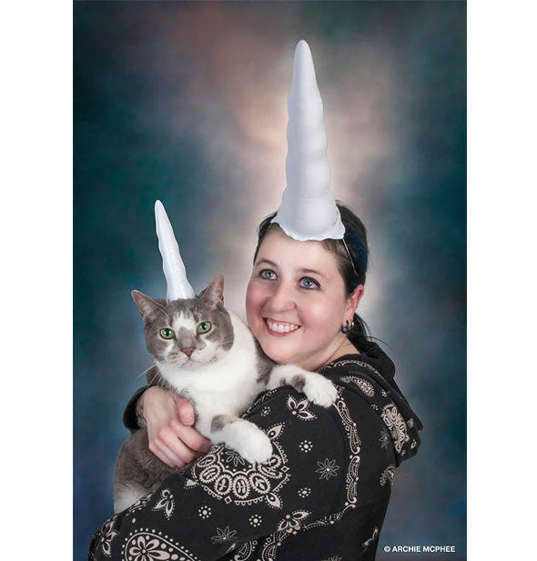 A smiling model poses with their cat, both wearing a white unicorn horn on their heads