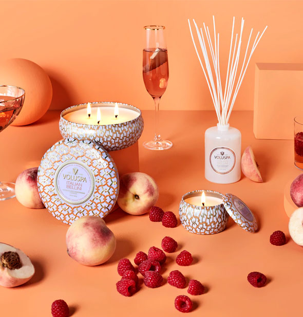 Voluspa candles and reed diffuser are staged on an orange background with raspberries, peaches, and champagne glasses