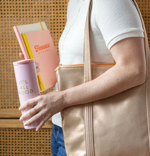 Model wearing a gold shoulder bag holds an It's All Good pink drink tumbler and notebooks