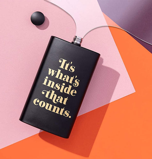 "It's what's inside that counts" matte finish black flask with cap removed is set on its side on a colorful surface