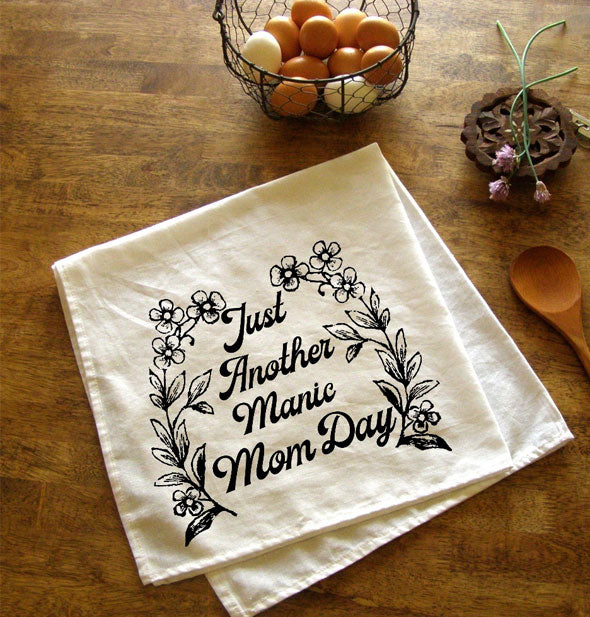 White towel with floral design and the saying, "Just Another Manic Mom Day" lays folded on a wooden tabletop with other kitchen accoutrements