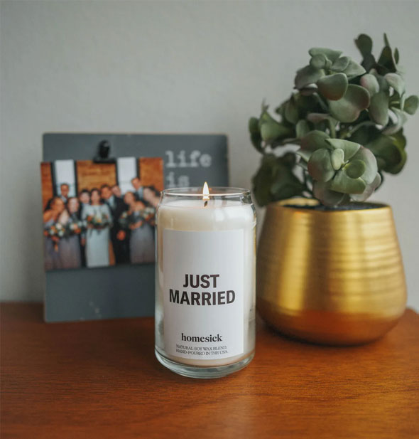A lit Just Married candle rests on a wooden tabletop with houseplant and group photo in the background