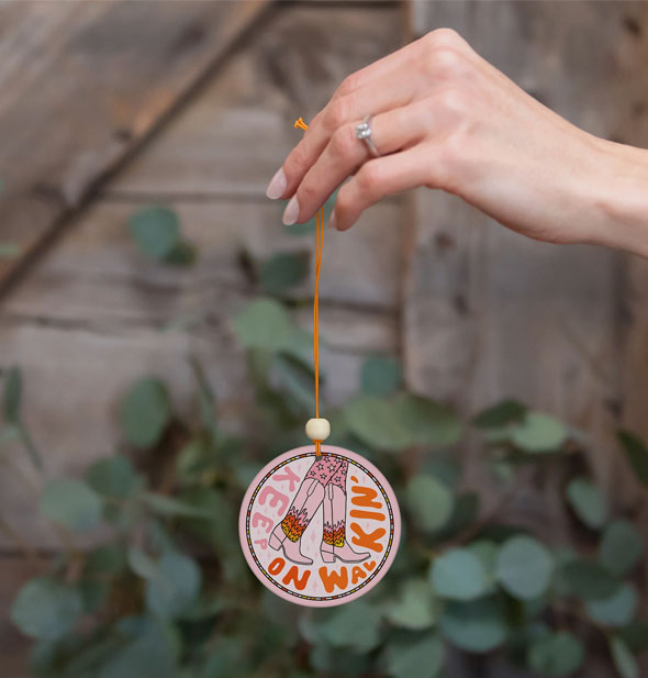 Model's hand holds a Keep On Walkin' car air freshener by its orange string against a botanical and wood backdrop