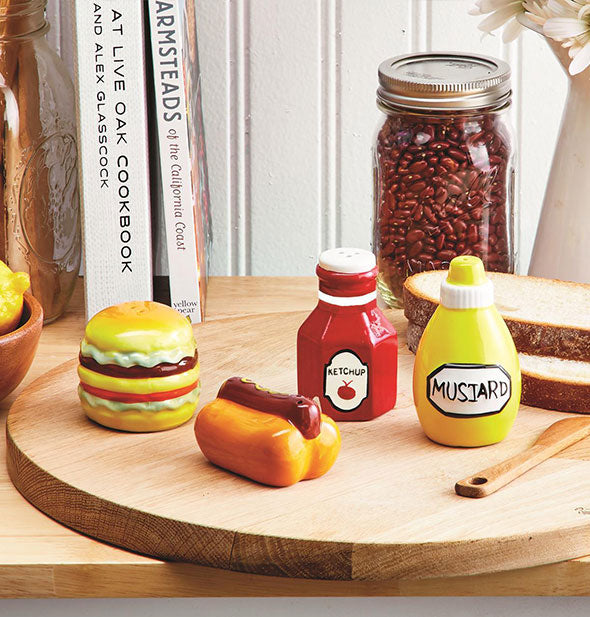 Hamburger, hot dog, ketchup, and mustard salt and pepper shakers are staged with jar of beans, cookbooks, and bread slices on a round wooden cutting board