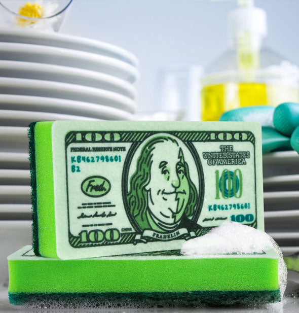 Two green hundred dollar bill dish sponges on a countertop with soap suds