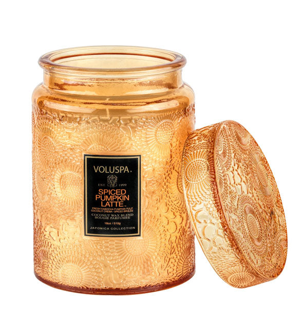 Burning candle in an orange embossed glass jar container with black label and matching lid set to the side.