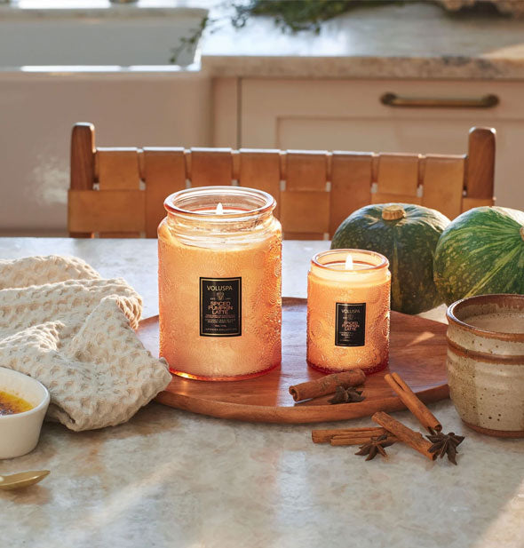 Small and large orange embossed glass jar candles on a kitchen tabletop with cinnamon sticks, star anise, and other accoutrements