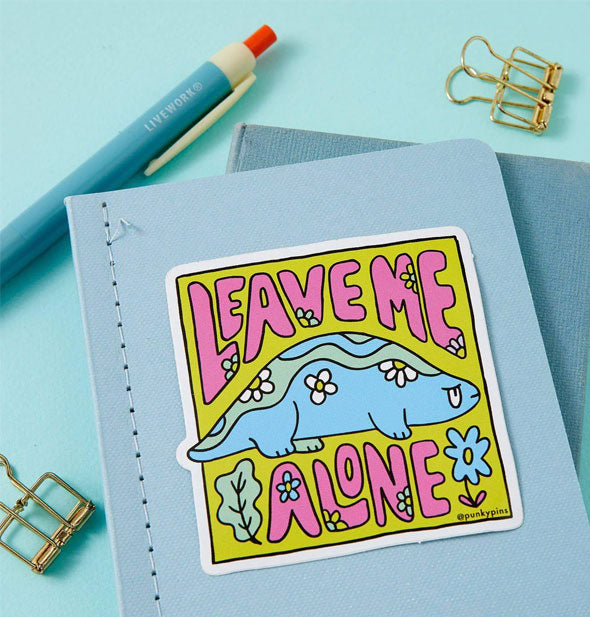 Leave Me Alone dinosaur sticker on a blue notebook cover with pen and paperclips surrounding it