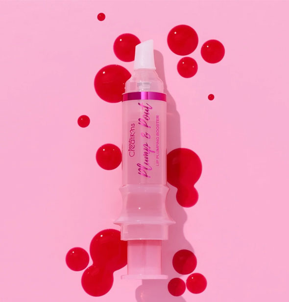 A syringe-shaped tube of Beauty Creations Plump & Pout lip gloss surrounded by bright reddish-pink droplets of product