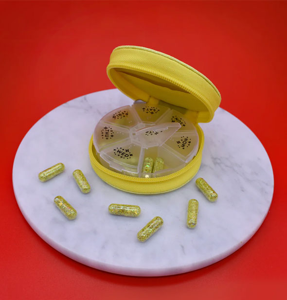 A round yellow pill case with plastic seven-day insert holding and surrounded by several yellow glitter capsules rests on a round marble disc on a red surface