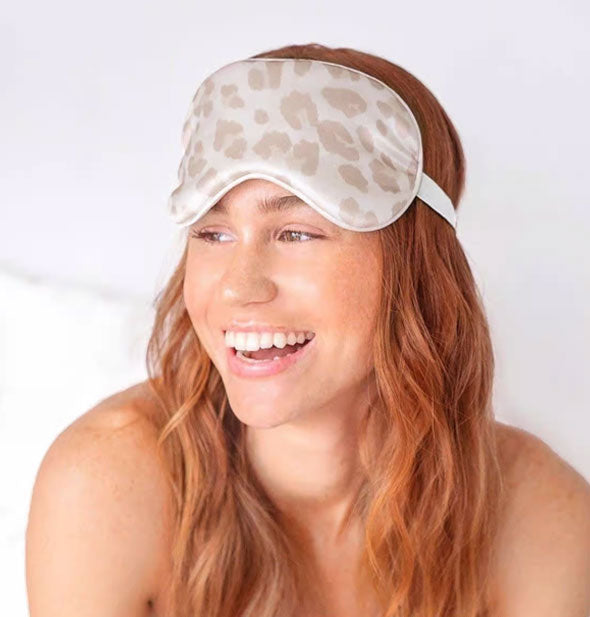 Smiling model wears a light brown and white leopard print sleep mask pushed up over eyes