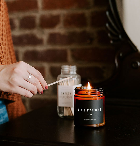 A model holds a match over a lit Let's Stay Home amber glass jar candle