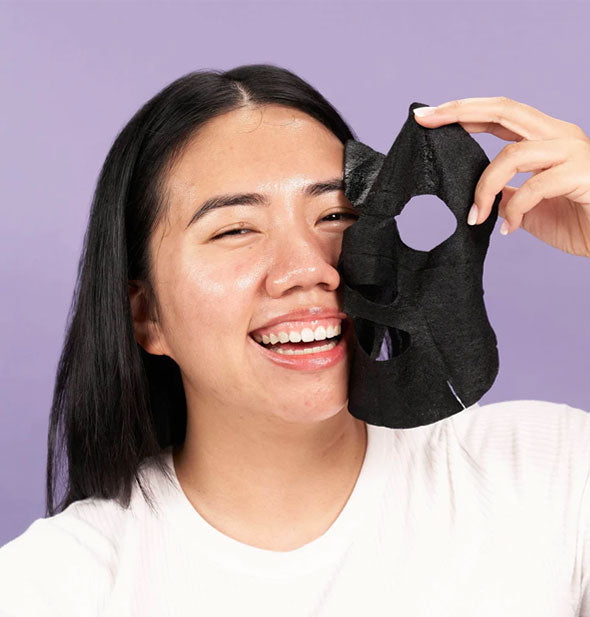 Smiling model pulls a black sheet mask away from face