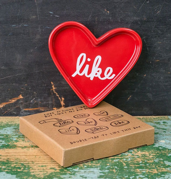 Red heart-shaped "Like" dish sits on top of a cardboard gift box on a distressed wooden backdrop