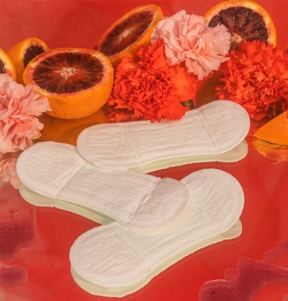 Three pantyliners on a red surface with red and pink flowers and halves of blood orange