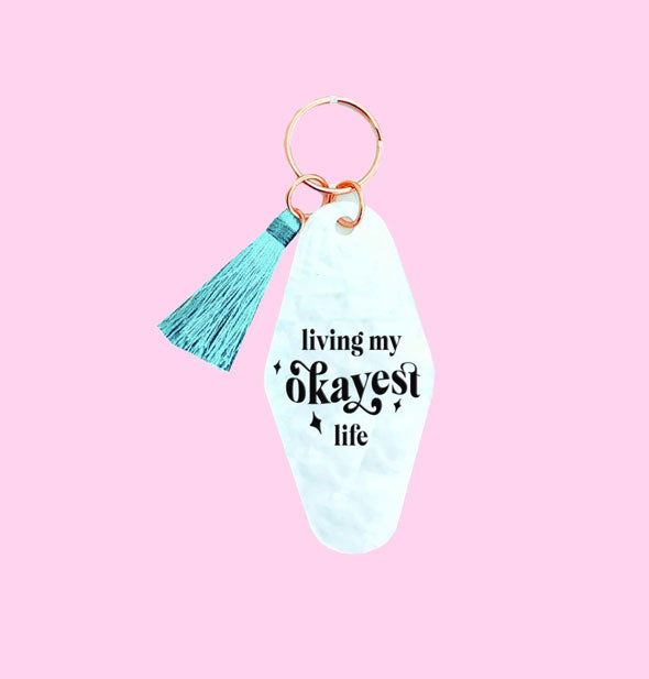 Swirled white acrylic keychain with teal tassel attached says, "Living My Okayest Life" in black lettering with star accents
