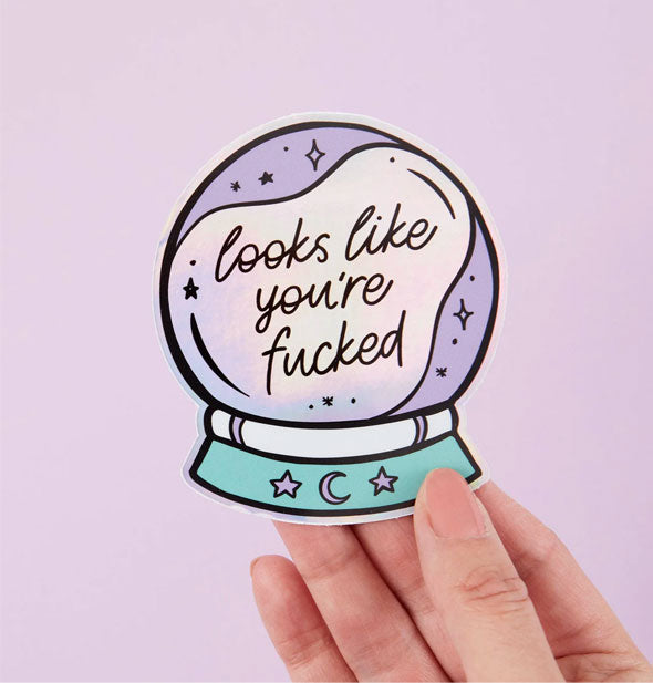 Model's hand holds sticker designed to resemble a crystal ball that says, "Looks like you're fucked" in black script surrounded by celestial accents