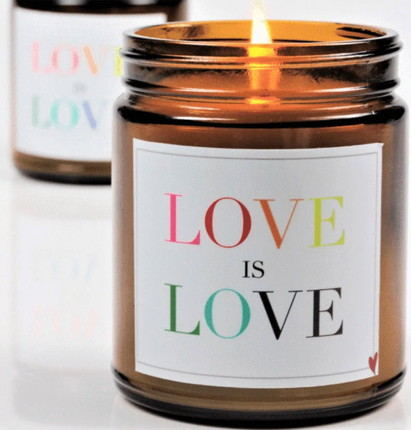 Lit amber glass jar candle with white label that says, "Love Is Love" in multicolored lettering
