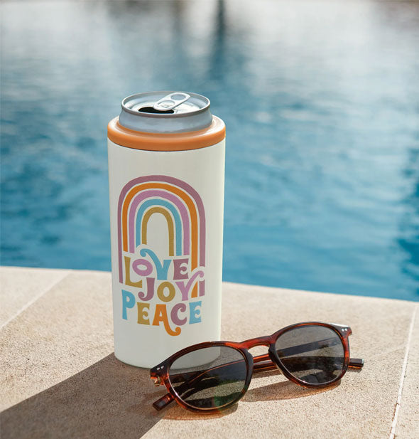 Love Joy Peace rainbow slim can cooler holds an opened can next to a pair of sunglasses at the edge of a swimming pool